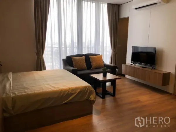 Condominium for rent in Khlong Toei, Bangkok - Living area with floor-to-ceiling curtains, sofa, TV console and bed on wood flooring.