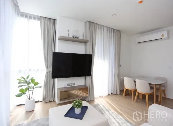 Condominium for rent or sale in Watthana, Bangkok - Sunlit lounge with wall-mounted TV, dining nook, air conditioner, sheer curtains and a potted plant.
