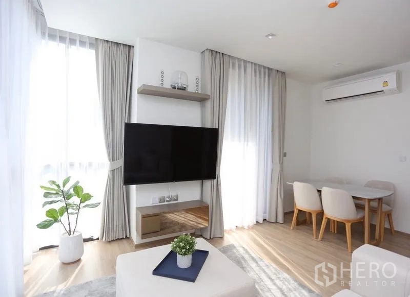 Condominium for rent or sale in Watthana, Bangkok - Sunlit lounge with wall-mounted TV, dining nook, air conditioner, sheer curtains and a potted plant.