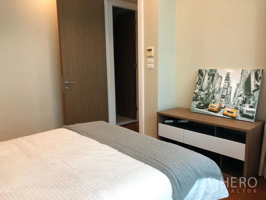 Condominium for rent or sale in Khlong Toei, Bangkok - Bedroom view toward doorway with low dresser and framed city artwork.