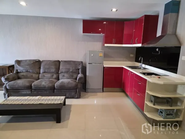 公寓 出租在Khlong Toei, Bangkok - 开放式客厅配灰色沙发与红色L形厨房，含炉灶与冰箱。