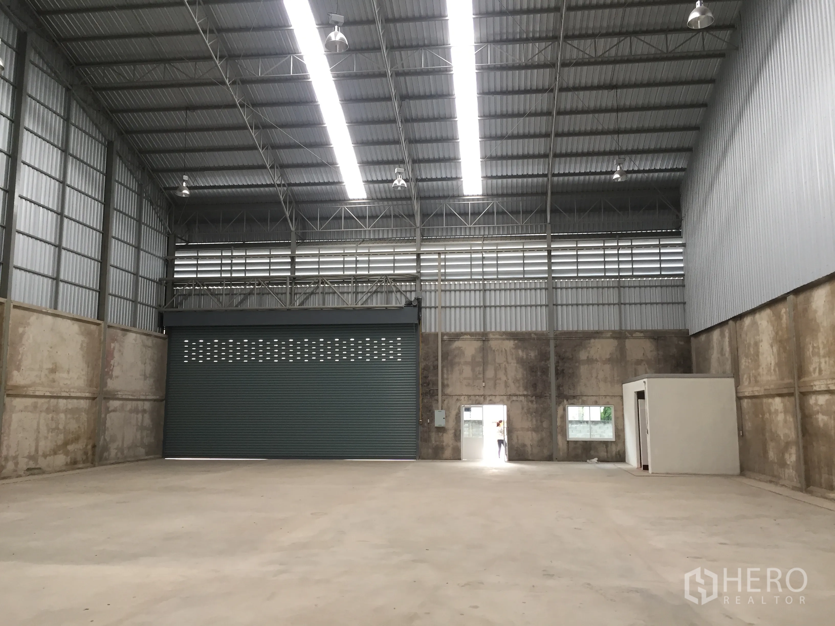 Warehouse for rent in Lam Luk Ka, Pathum Thani - Open-plan warehouse showing electric roller shutter, small office room and rear exit doors.
