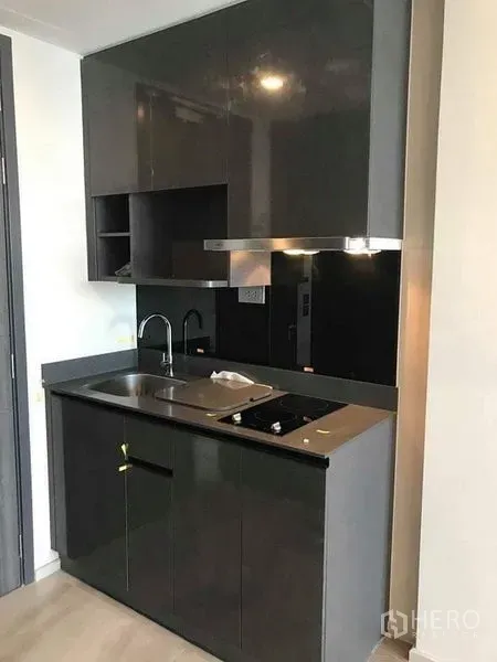 Condominium for rent in Watthana, Bangkok - Compact modern kitchenette with glossy cabinets, sink and electric cooktop.