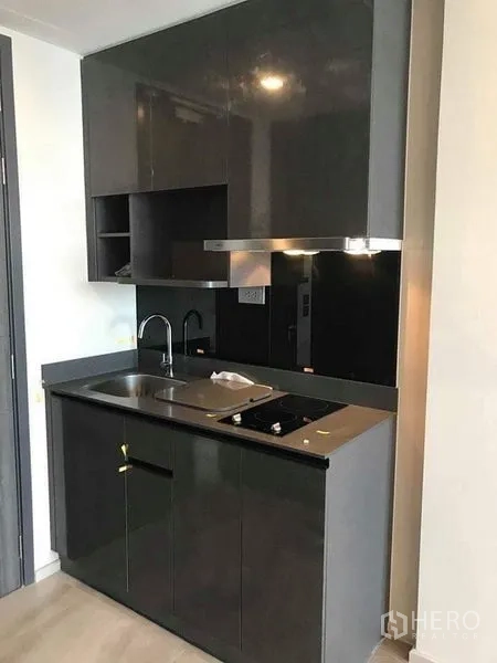 Condominium for rent in Watthana, Bangkok - Compact modern kitchenette with glossy cabinets, sink and electric cooktop.