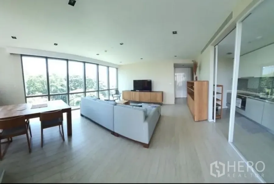 公寓 出租或出售在Watthana, Bangkok - 开放式客餐厅，落地窗、L形沙发与电视柜，旁连玻璃隔断厨房，位于瓦塔那公寓