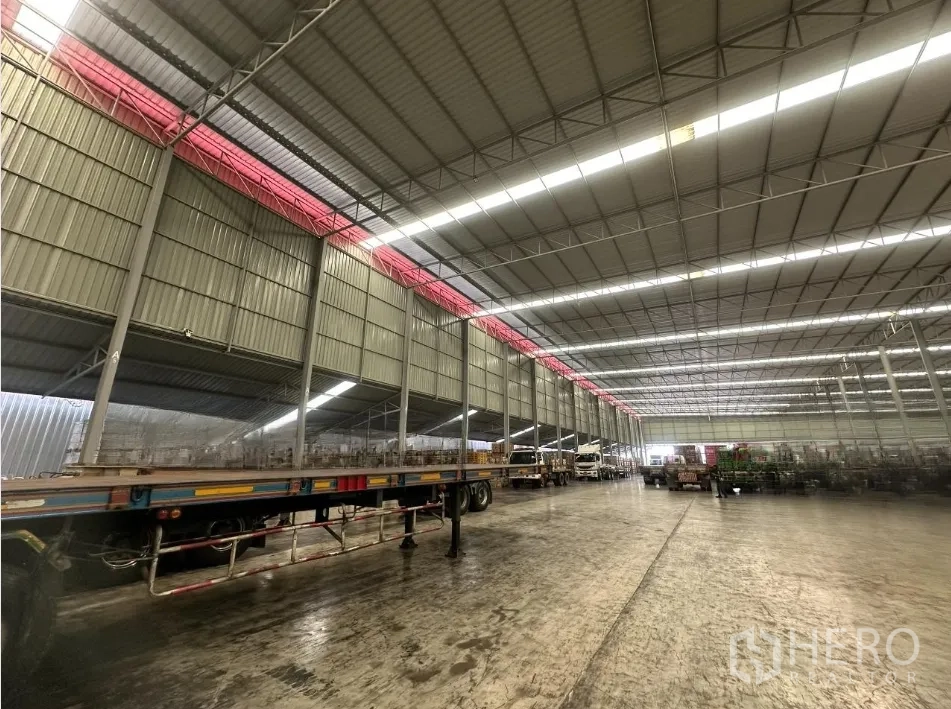 Warehouse for rent in Bang Yai, Nonthaburi - Large warehouse interior with high-clearance steel roof, skylight panels, flatbed trailers and concrete floor.