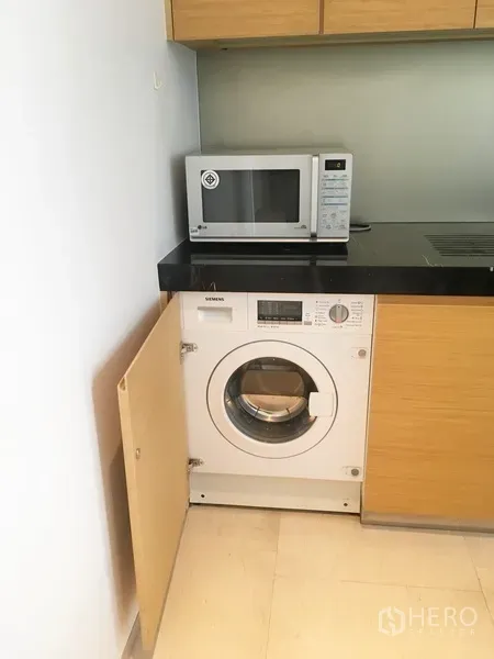 Condominium for rent in Bang Rak, Bangkok - Compact kitchen with black countertop, microwave, and built-in front-load washer behind cabinet door.