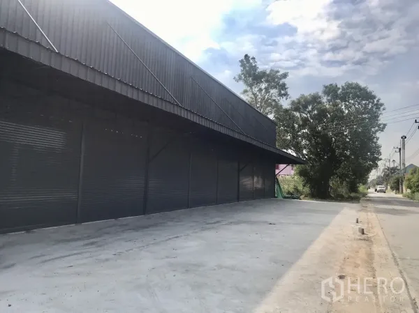 Warehouse for rent in Lam Luk Ka, Pathum Thani - Long warehouse facade under a canopy with ample driveway and roadside trees.