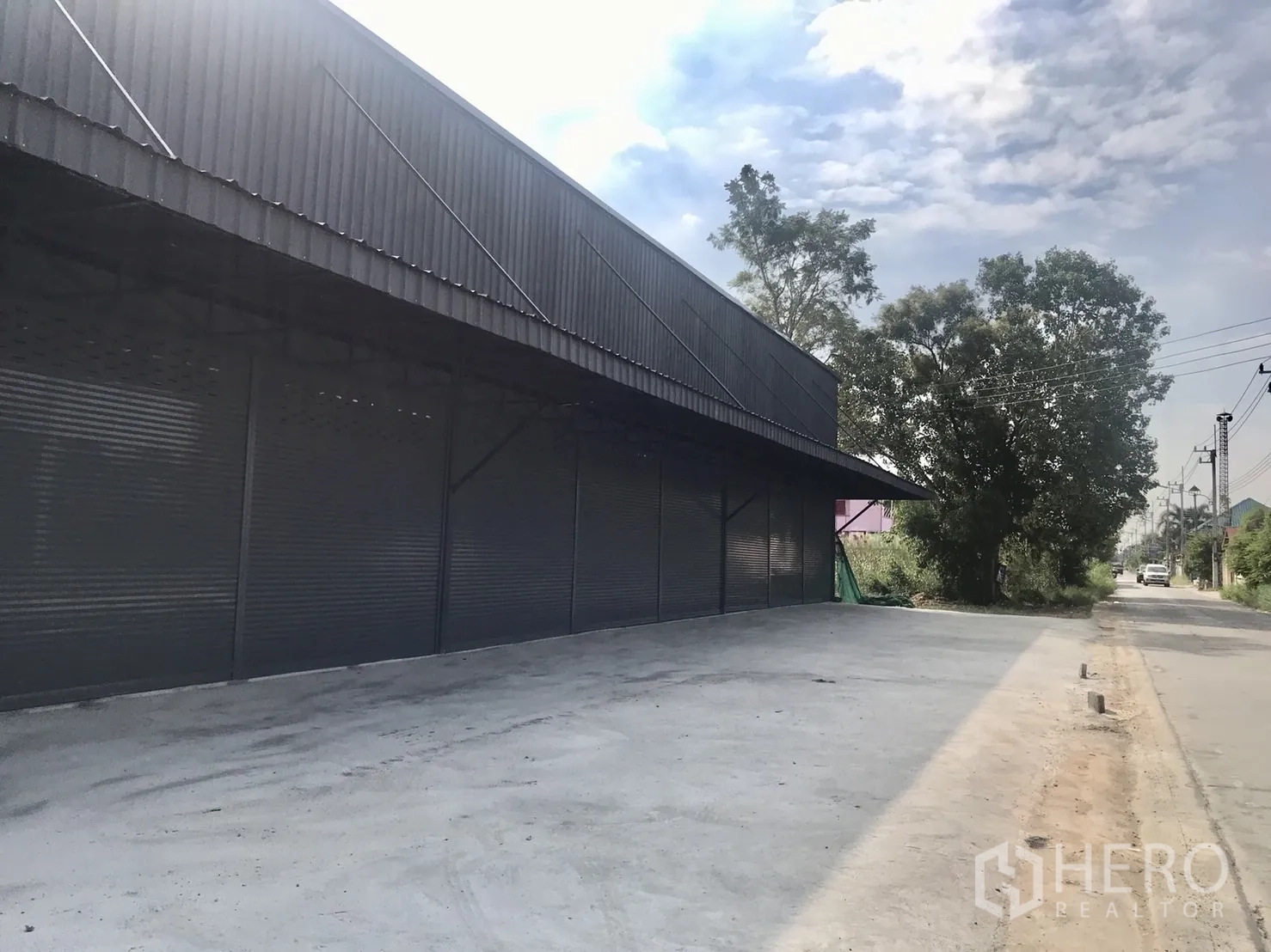 Warehouse for rent in Lam Luk Ka, Pathum Thani - Long warehouse facade under a canopy with ample driveway and roadside trees.