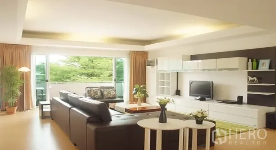 Apartment for rent in Khlong Toei, Bangkok - Bright living room with L-shaped sofa, TV wall and sliding doors opening to a green balcony view