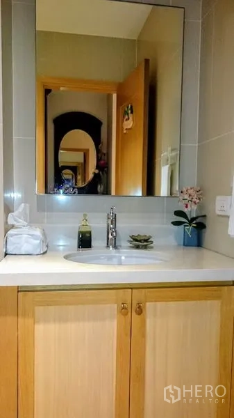 Condominium for rent in Khlong Toei, Bangkok - Bathroom vanity with large mirror, under-sink cabinet and small flower décor.