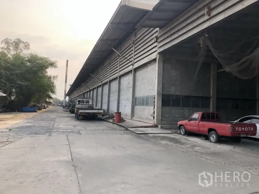 Factory for rent in Bang Bua Thong, Nonthaburi - Long factory exterior with covered loading bays and trucks on a wide concrete drive.