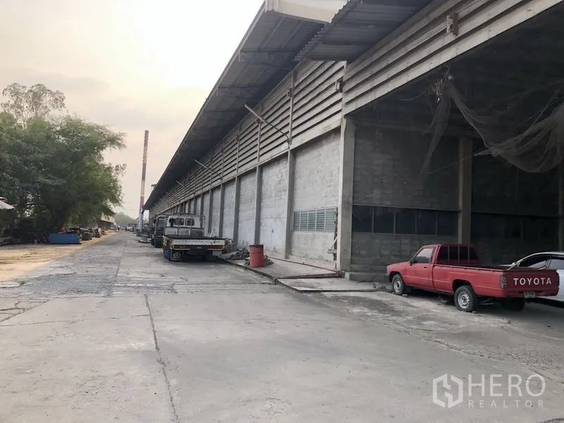 Factory for rent in Bang Bua Thong, Nonthaburi - Long factory exterior with covered loading bays and trucks on a wide concrete drive.