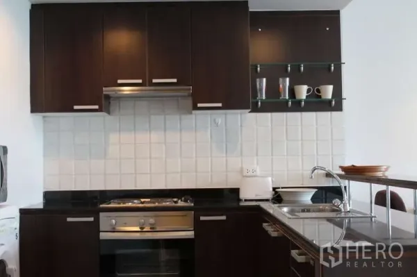 Condominium for rent in Sathon, Bangkok - Modern kitchen with dark wood cabinetry, stovetop and oven, white tile backsplash, and glass shelves with cups.