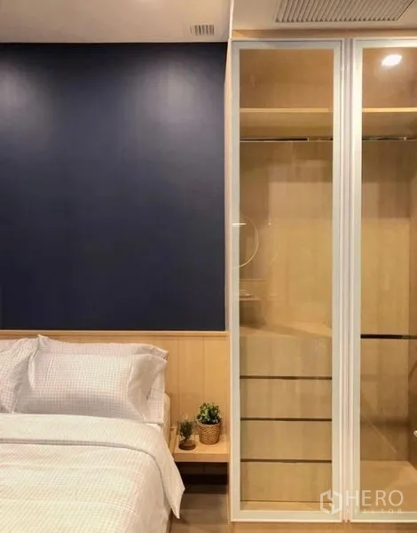 Condominium for rent in Watthana, Bangkok - Bedroom corner with white bedding beside a glass sliding-door wardrobe and a navy accent wall.