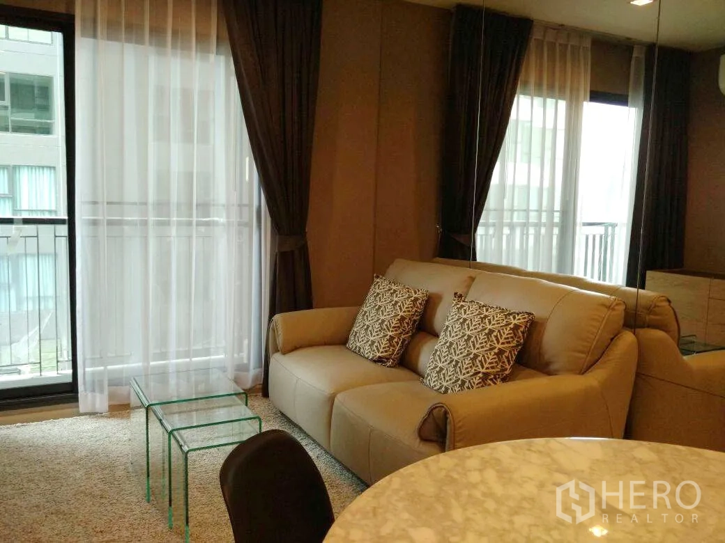 Condominium for rent in Khlong Toei, Bangkok - Bright living room with beige sofa, floor-to-ceiling windows, sheer curtains, and balcony outlook.