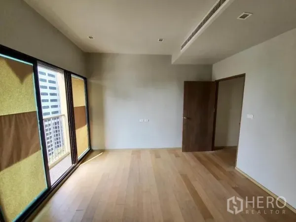 公寓 出售在Ratchathewi, Bangkok - 明亮的无家具起居区，浅色木地板，落地推拉门通向小阳台，可见城市建筑景观。