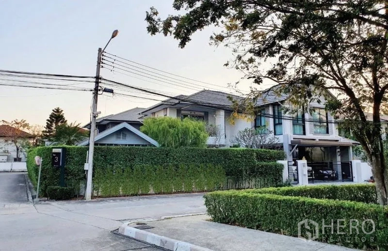 独立式住宅 出售在Lam Luk Ka, Pathum Thani - 转角地块的双层独栋别墅，绿色树篱围合，带车棚与林荫街道景观。