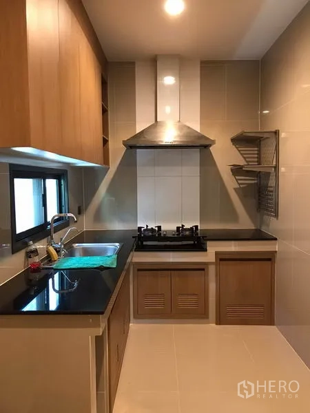 Detached House for rent in Bang Kapi, Bangkok - Enclosed kitchen with black countertops, gas hob, stainless hood and wood cabinetry.