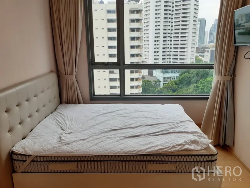 Condominium for rent in Watthana, Bangkok - Cozy bedroom with large window, upholstered headboard, and skyline with greenery views.