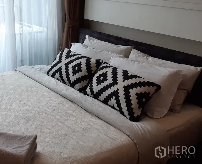 Condominium for rent in Khlong Toei, Bangkok - Bedroom with large bed, monochrome patterned cushions and balcony light through curtains.