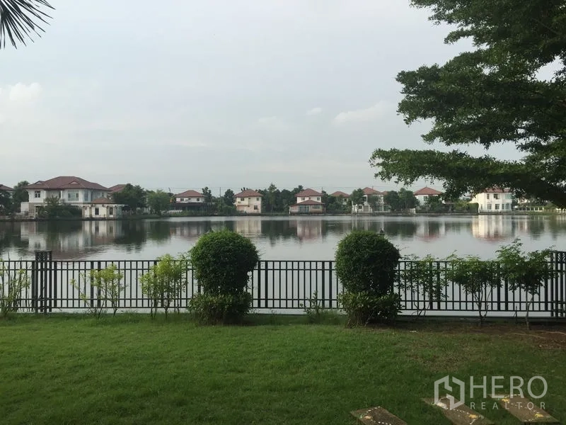 Detached House for rent or sale in Lat Krabang, Bangkok - Private lawn with metal fence overlooking a calm lake and neighboring homes.