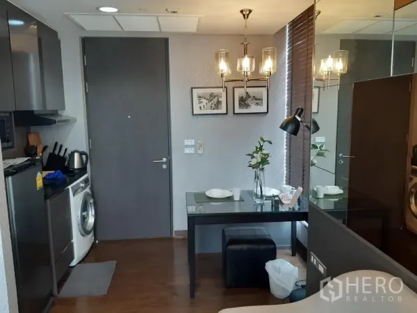 Condominium for rent in Khlong Toei, Bangkok - Compact kitchen-dining area with built-in cabinets, fridge, microwave, washing machine and a two-seat table under a chandelier and mirrored wall.