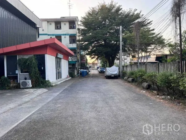 货栈 出租在Lat Phrao, Bangkok - 仓库外小巷景观，红檐办公楼、行道树与路边停车位。