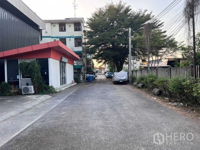 货栈 出租在Lat Phrao, Bangkok - 仓库外小巷景观，红檐办公楼、行道树与路边停车位。