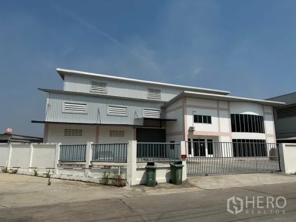 货栈 出租在Samut Prakan - 街景：带栅栏与大门的仓库园区，设停车与卷闸入口。