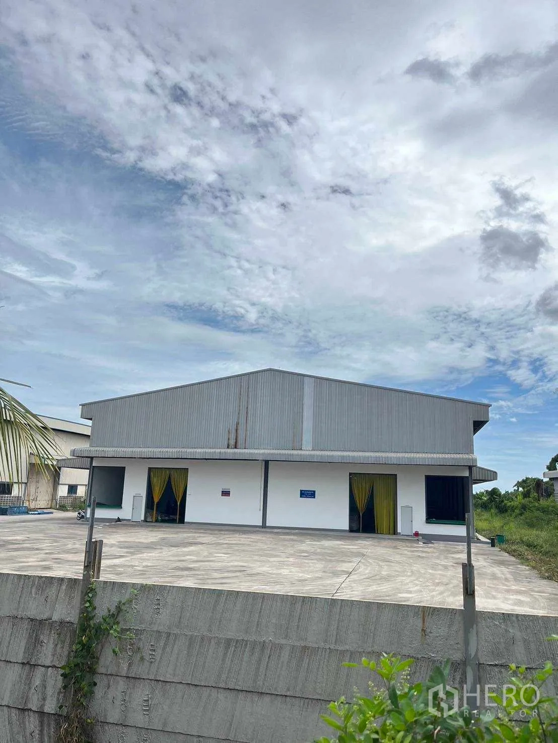 โรงงาน ขายในNikhom Phatthana, Rayong - ด้านหน้าโกดังมีชายคาบริเวณโหลดสินค้าและลานคอนกรีตยกสูงหลังรั้ว