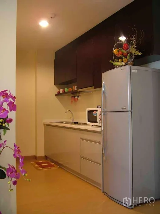 公寓 出租或出售在Watthana, Bangkok - 紧凑一字型厨房，深色吊柜、水槽、微波炉和高大银色冰箱。