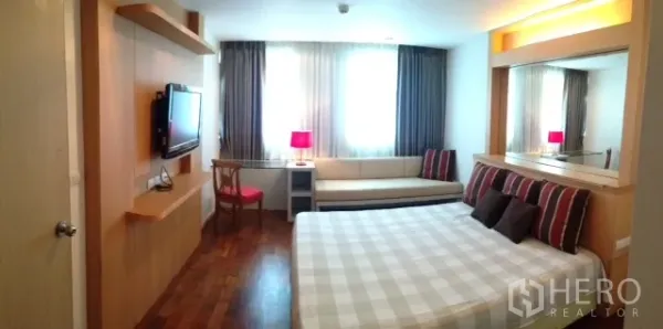Condominium for rent or sale in Khlong Toei, Bangkok - Bright bedroom with a king bed, wall-mounted TV, sofa by the window, wood flooring and a mirrored headboard.