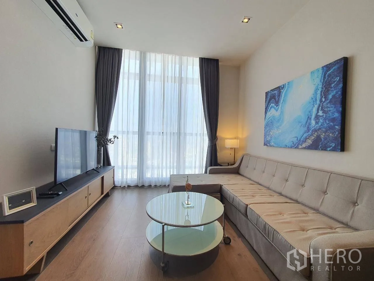 Condominium for rent in Khlong Toei, Bangkok - Living room with large TV console, wall art, and tall windows with sheer curtains.