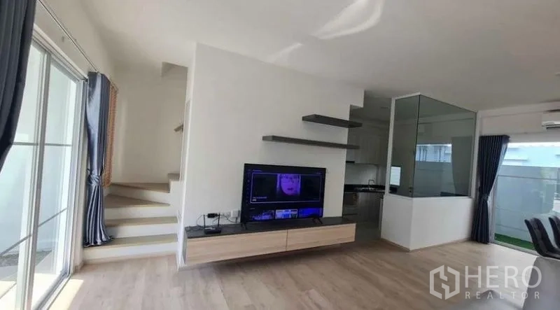 Townhouse for rent in Prawet, Bangkok - Living room with floating TV console, wall shelves and staircase to upper floor.