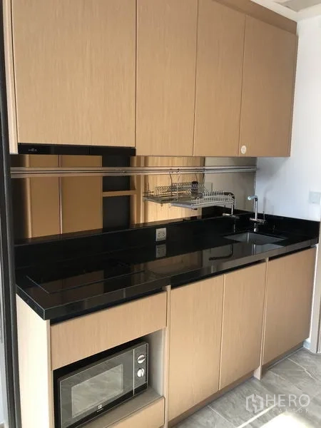 Condominium for rent in Watthana, Bangkok - Compact kitchenette with black stone countertop, sink, cooktop, microwave, and ample cabinets.