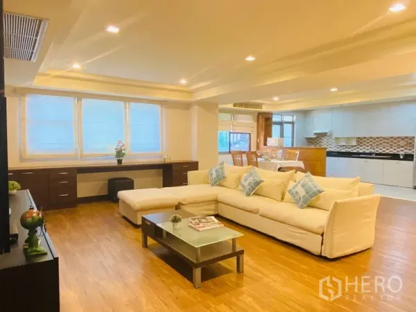 公寓 出租在Watthana, Bangkok - 开放式客厅，白色L型沙发、玻璃茶几与木地板，后方为带马赛克墙面的明亮厨房。