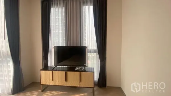 Condominium for rent - TV on wooden console between full-height windows with dark drapes and sheer curtains.
