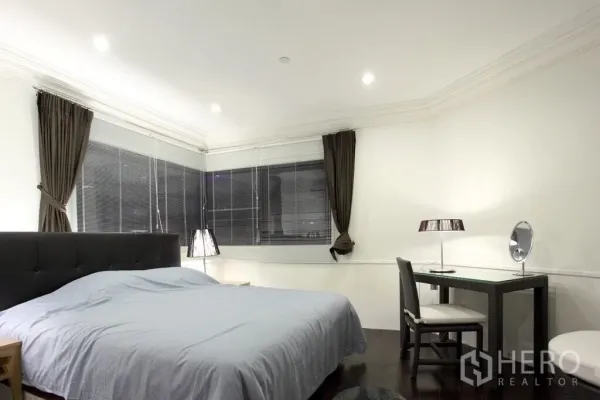 Condominium for rent in Sathon, Bangkok - Spacious bedroom with upholstered headboard, work desk, and corner windows with blinds and curtains.
