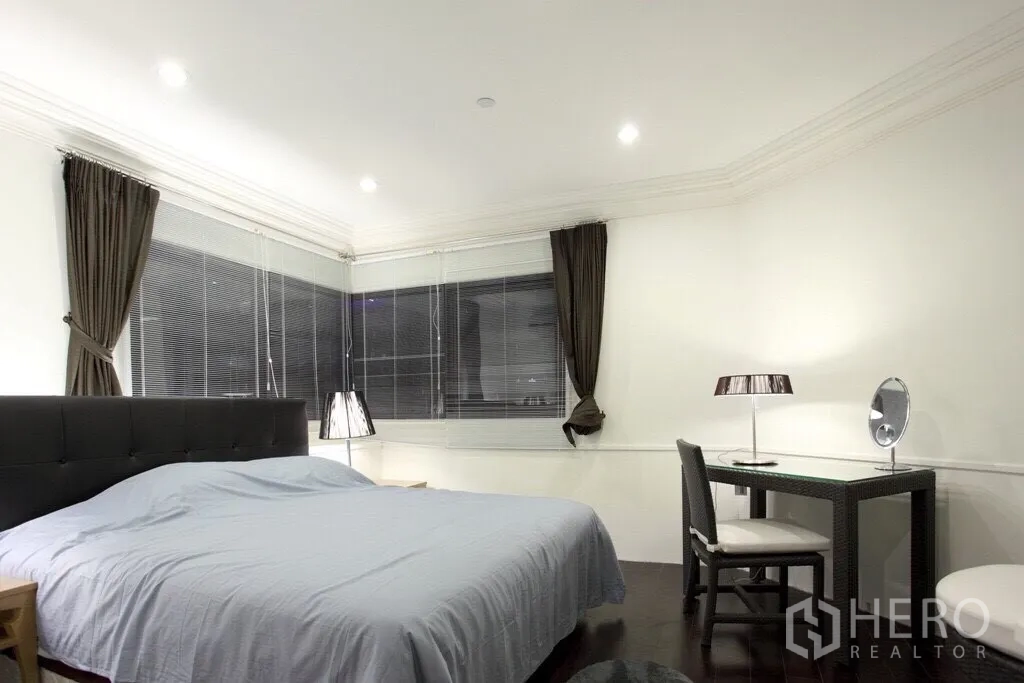 Condominium for rent in Sathon, Bangkok - Spacious bedroom with upholstered headboard, work desk, and corner windows with blinds and curtains.