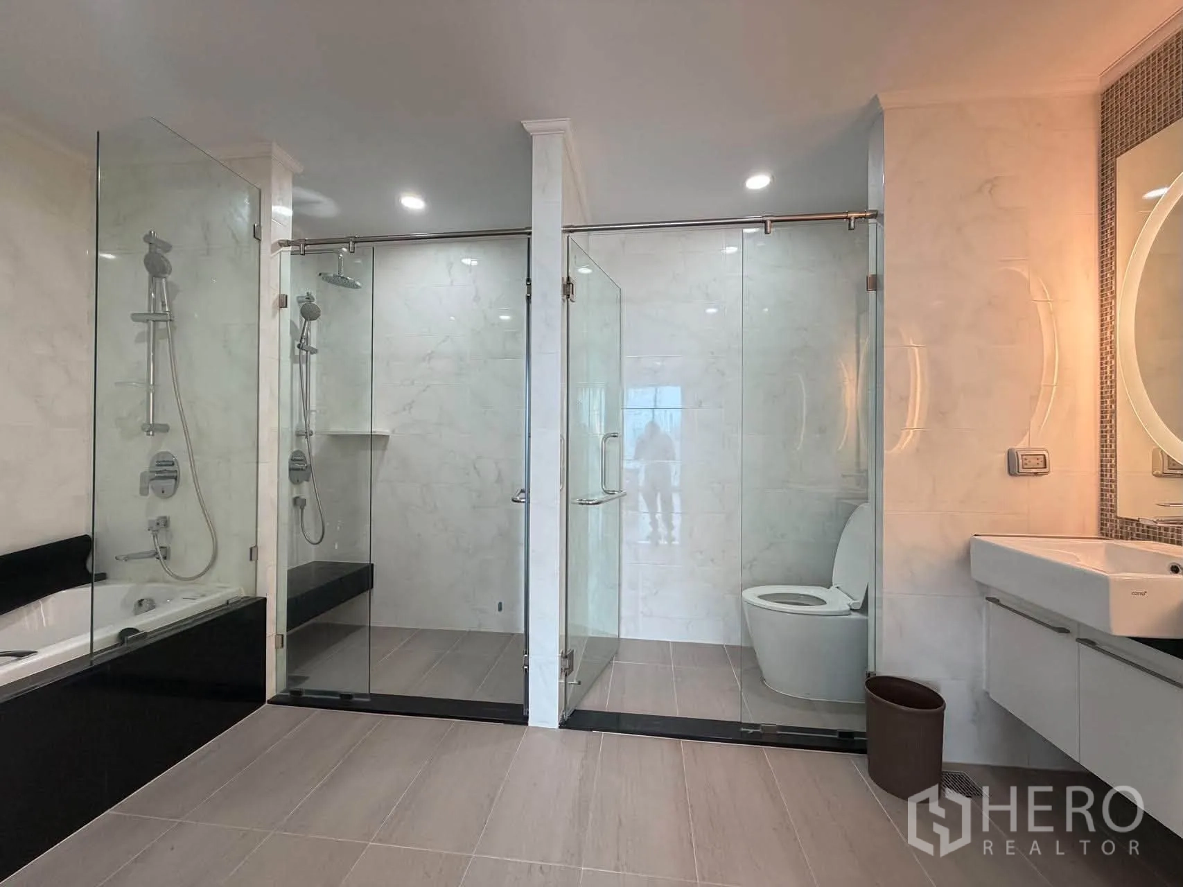 Condominium for sale in Watthana, Bangkok - Large bathroom with dual glass showers, vanity, and toilet under recessed lights.