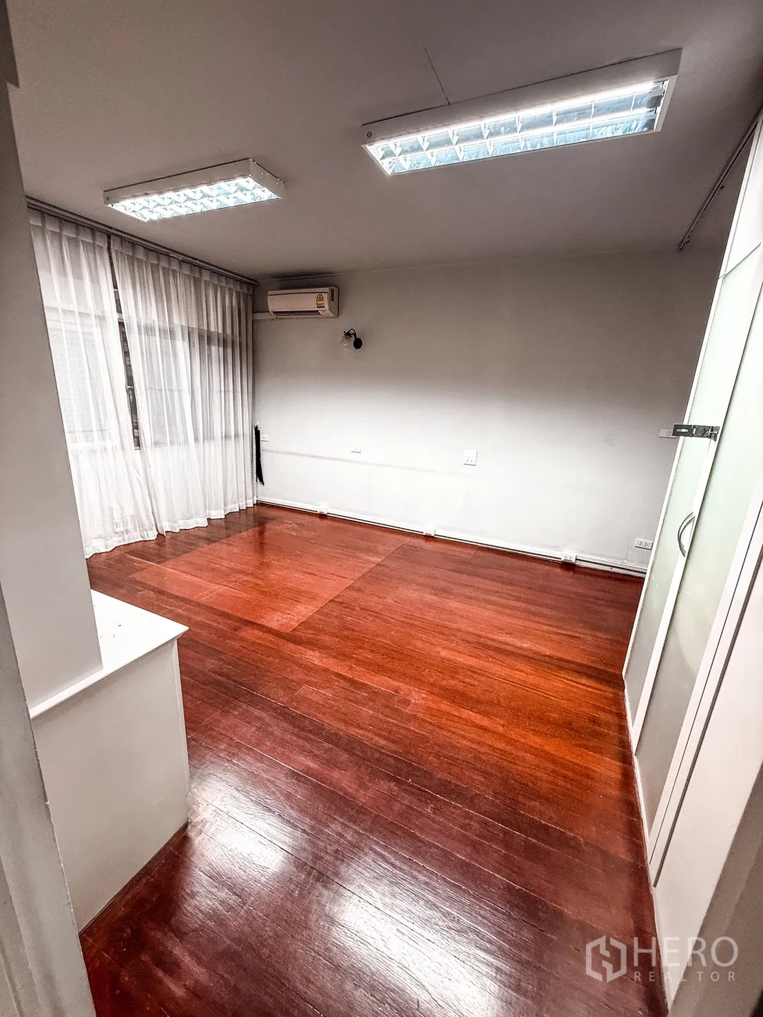Home Office for rent in Watthana, Bangkok - Bright office room with polished wooden floor, sheer curtains, fluorescent ceiling lights, and a wall-mounted air conditioner.
