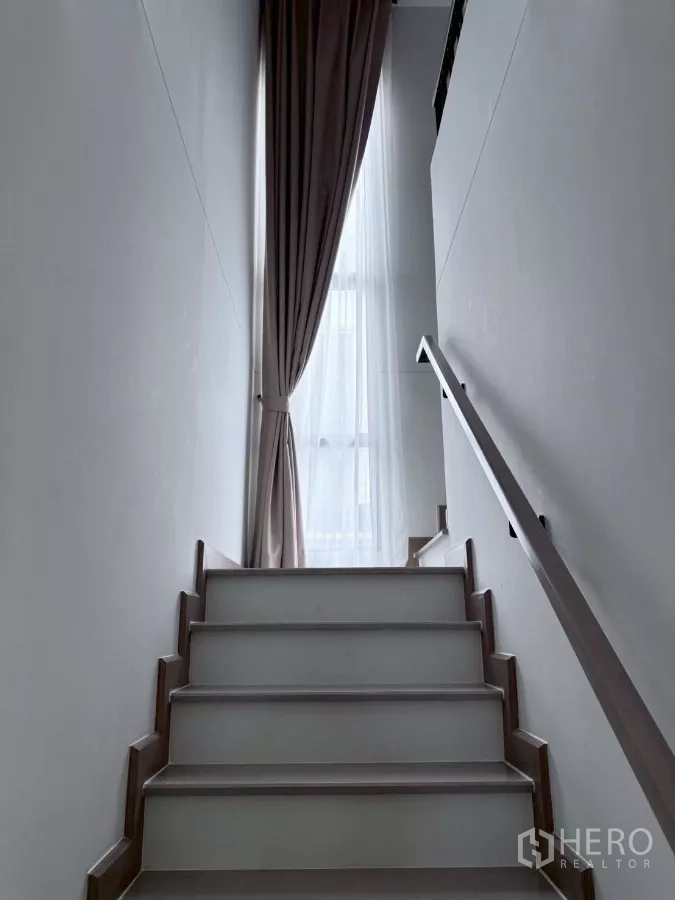 Detached House for rent in Lat Krabang, Bangkok - Staircase with handrail leading to upper floor, lit by tall window and drapes.