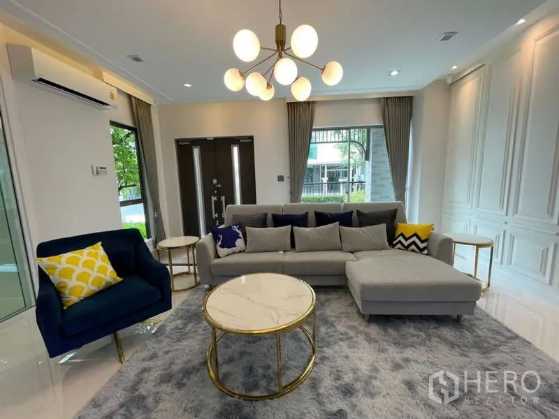 Detached House for rent or sale in Lat Krabang, Bangkok - Main living room with a gray sectional sofa, gold-accent coffee tables and a modern chandelier.