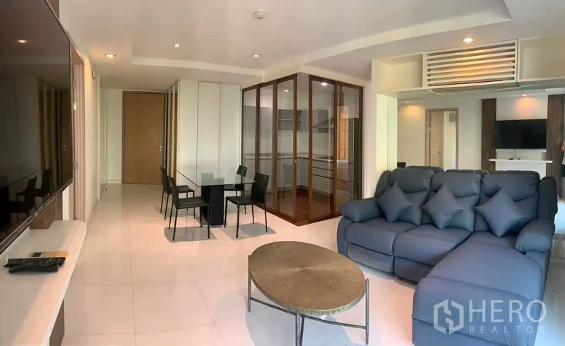 Condominium for rent or sale in Khlong Toei, Bangkok - Open-plan living with blue sofa, dining set, and a glass-walled kitchen.