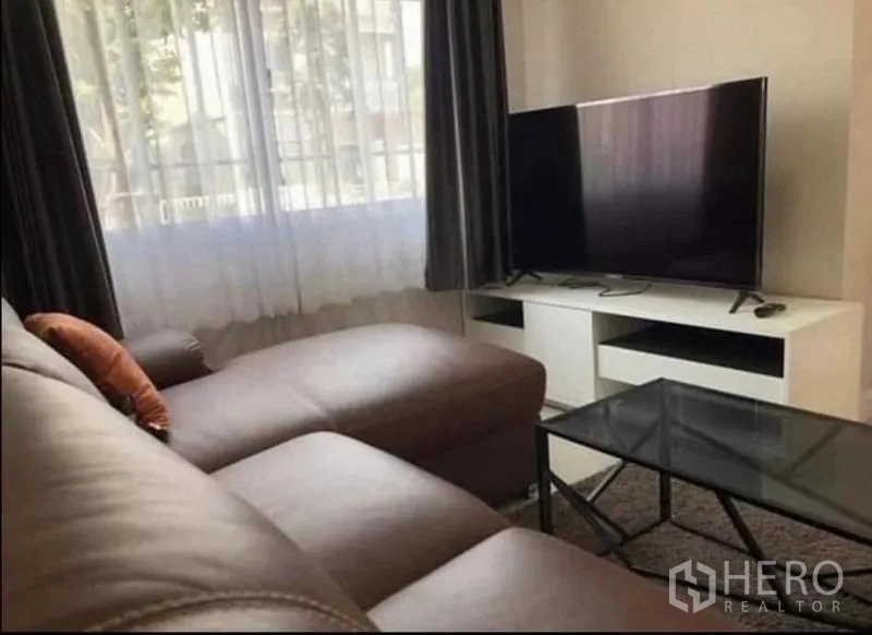 带办公室的家 出租在Samut Prakan - 采光充足的客厅，棕色L形皮沙发、玻璃茶几与电视柜，旁有窗帘大窗。