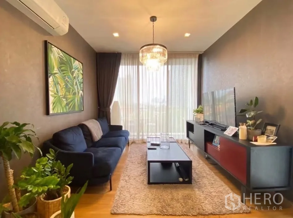Condominium for rent in Watthana, Bangkok - Inviting living room with navy sofa, chandelier, plants, and sheer floor-to-ceiling curtains.