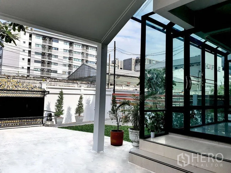 独立式住宅 出租在Phra Khanong, Bangkok - 覆顶露台与玻璃墙，连接花园与车道，可见城市楼景。