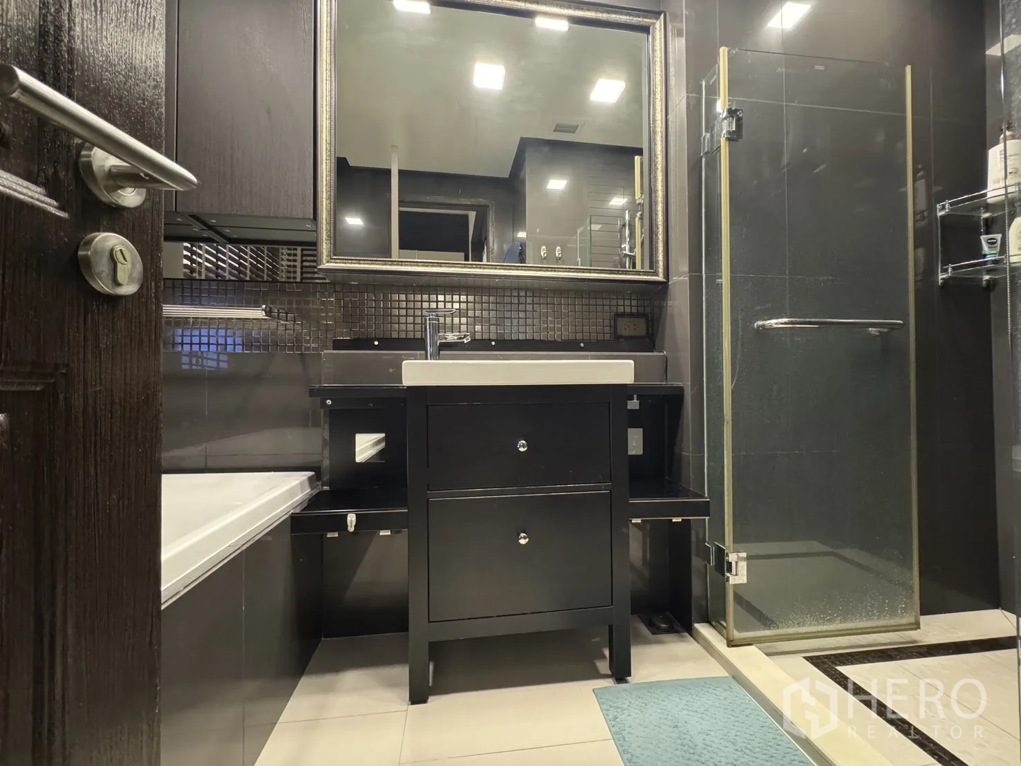 Condominium for rent or sale in Khlong Toei, Bangkok - Bathroom vanity with framed mirror and glass shower door in dark finishes.