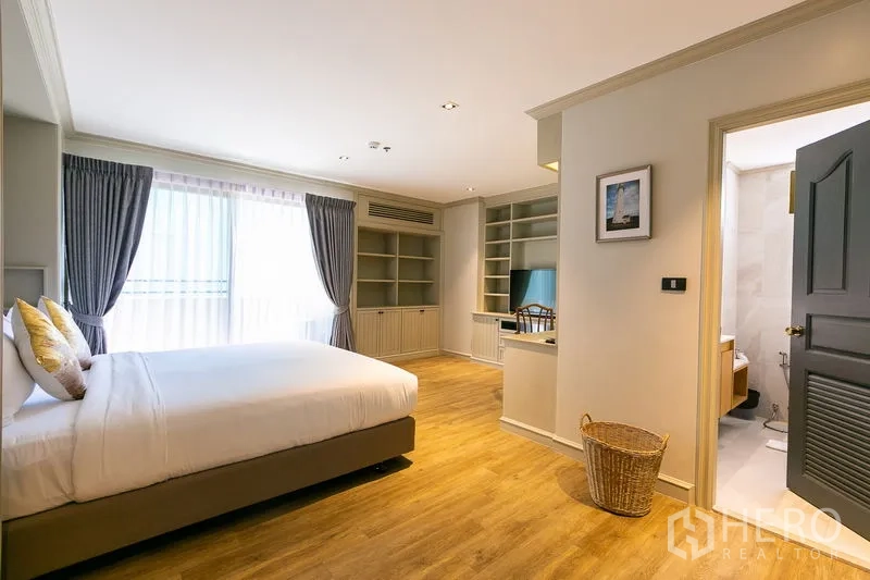 Apartment for rent in Watthana, Bangkok - Primary bedroom with large window, built-in shelving and workspace, with ensuite bathroom entrance.